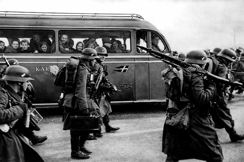 Nemeckí vojaci s výzbrojou počas obsadenia Dánska v roku 1940, civilisti sledujú situáciu z autobusu