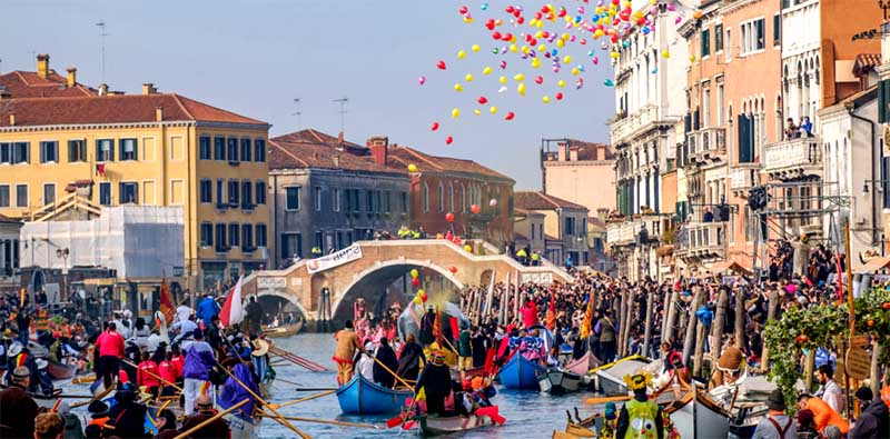 Benátsky karneval na hlavnom kanáli s farebnými loďami, balónmi a historickým mostom