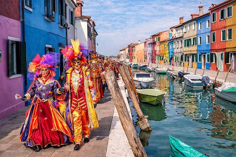 Sprievod Benátskeho karnevalu v pestrofarebných maskách pozdĺž kanála na ostrove Burano