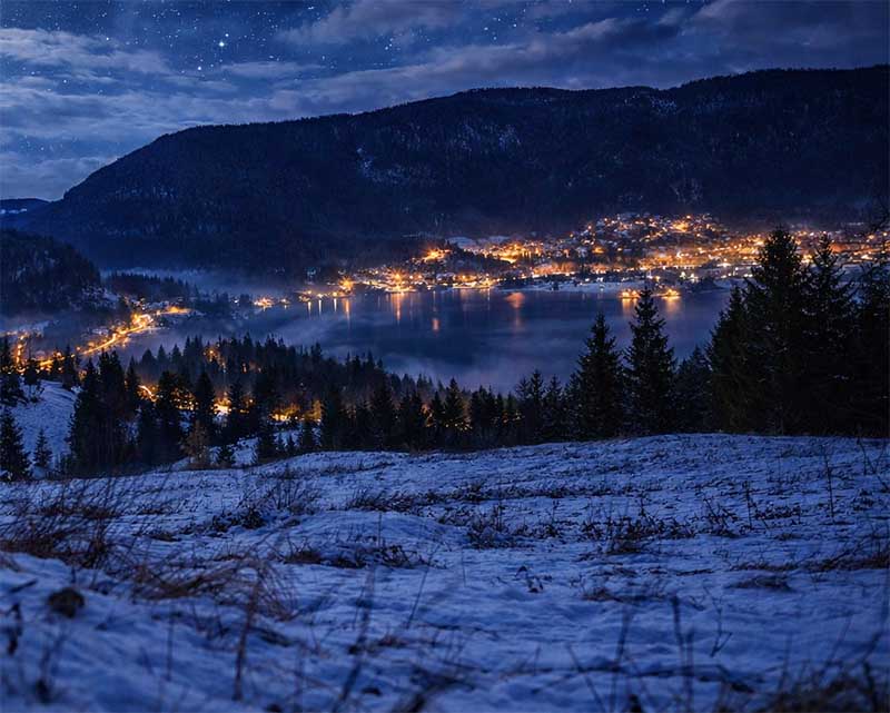 Nočná zimná krajina s osvetlenou obcou pri jazere pod horami, mesačné svetlo a hviezdna obloha